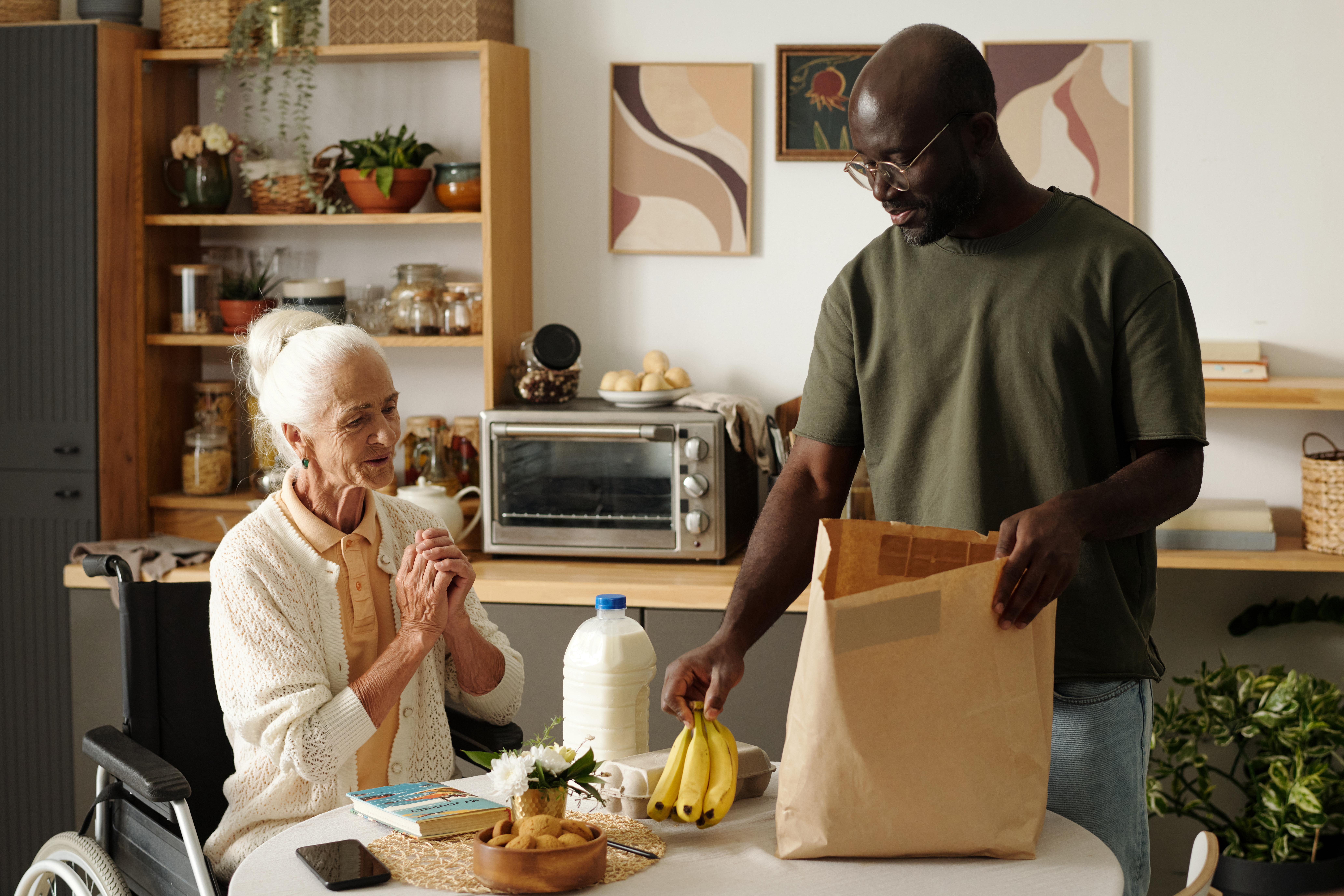 Senior Caucasian woman sitting in wheelchair smiling while Black man unloading groceries including bananas and milk onto kitchen table, both interacting in home setting