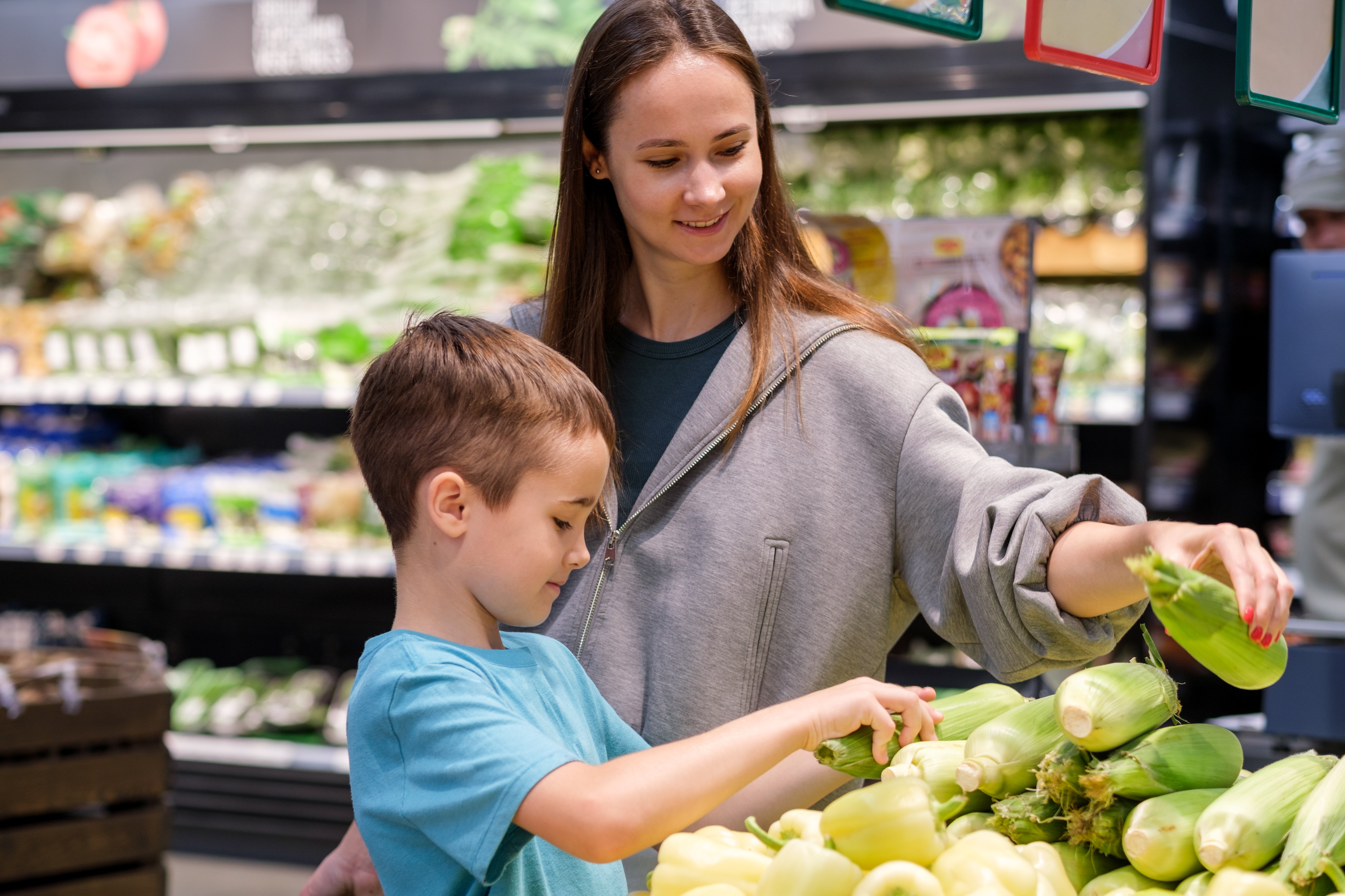 Mother and child shop for fresh corn in grocery aisle. Young boy looks up with curiosity, smiling at his mother. She gently holds a bright green corn cob, guiding his choice